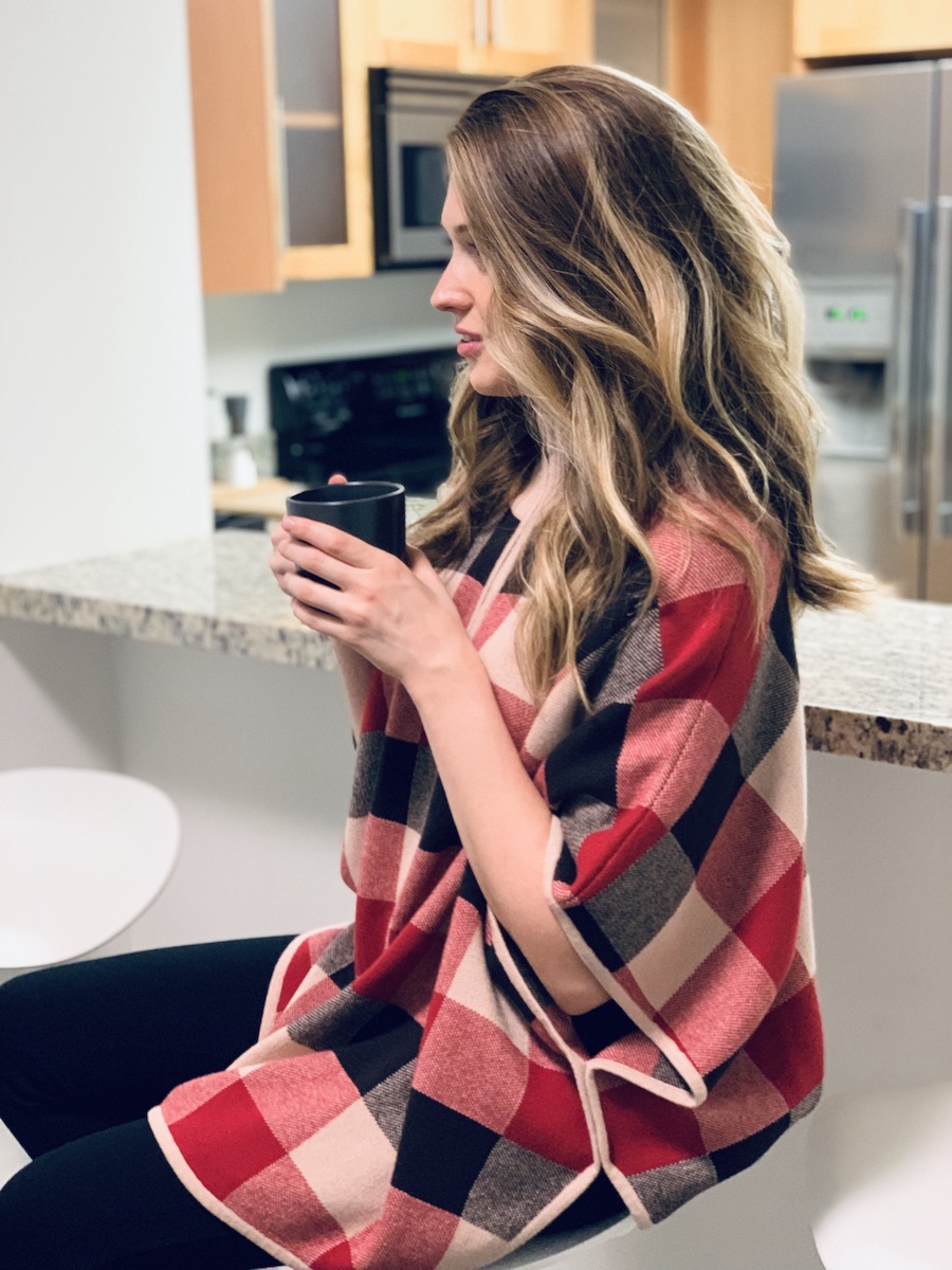 Woman sitting at a counter holding a cup of coffee wearing a Stylevate Plaid Mock Neck Cape